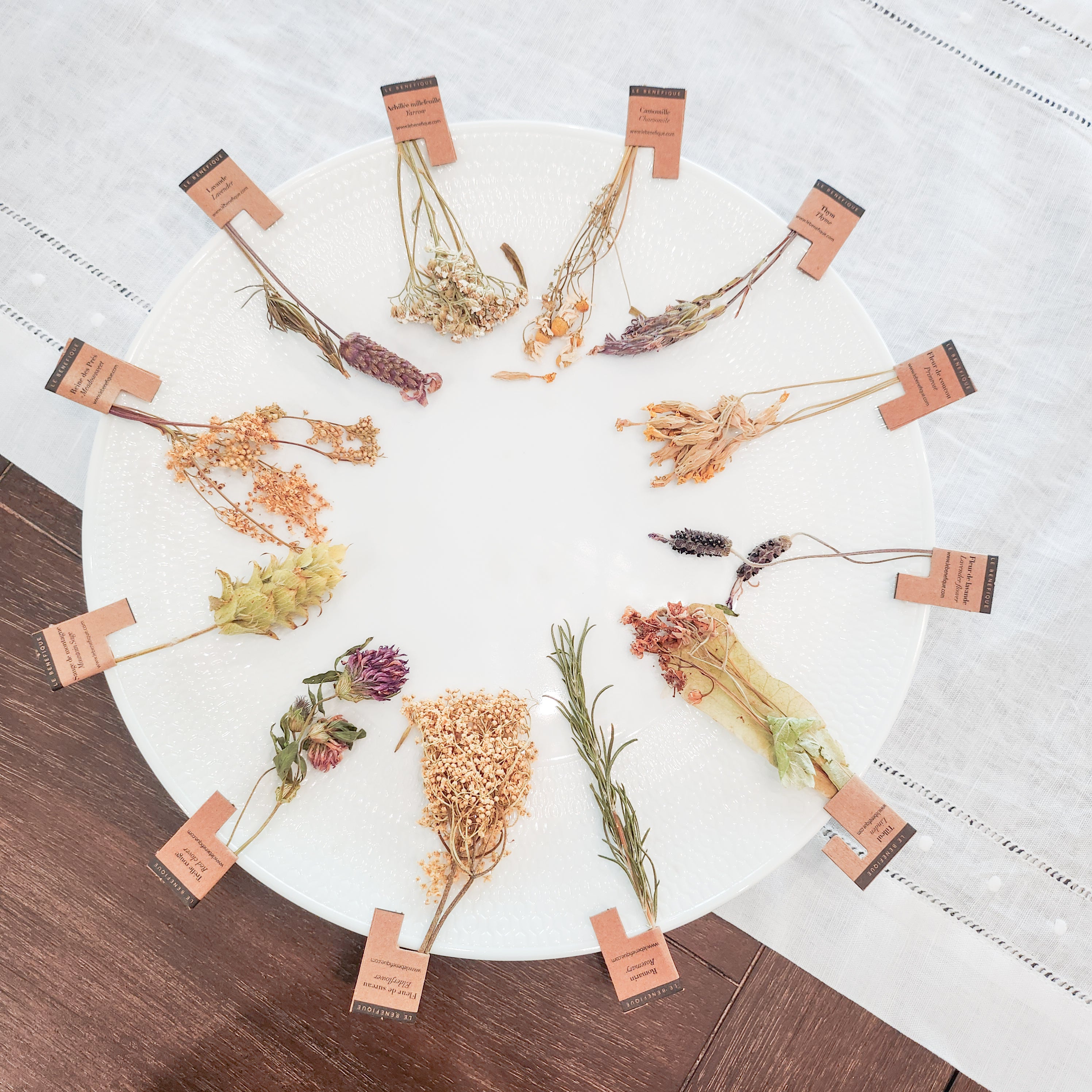 Overhead view of a white plate displaying 12 different dried flowers with small brown labels attached, on a wood surface with a white tablecloth in the background.