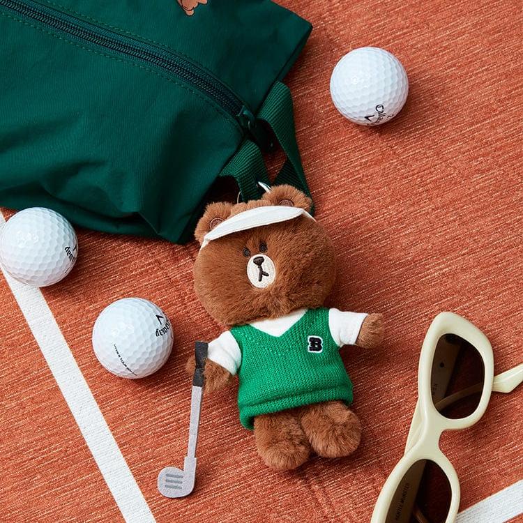 Brown teddy bear golfer with green vest, golf club, white visor, golf balls, green bag, and cream sunglasses on an orange surface with a white stripe.
