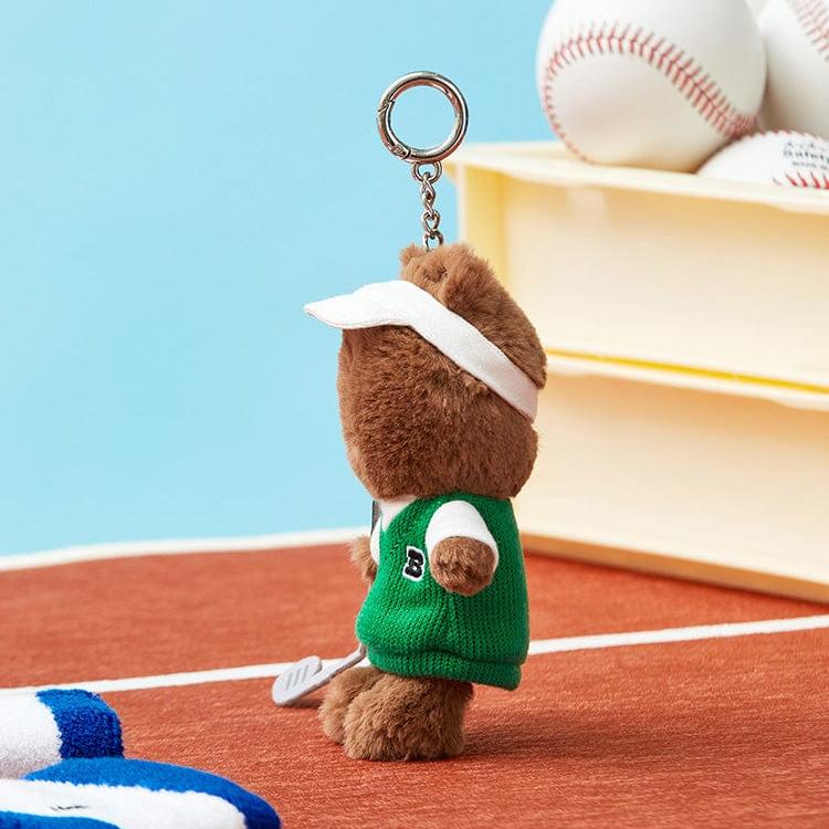 Side view of a brown teddy bear keychain wearing a white visor and green sweater with a white "B" on a red surface.