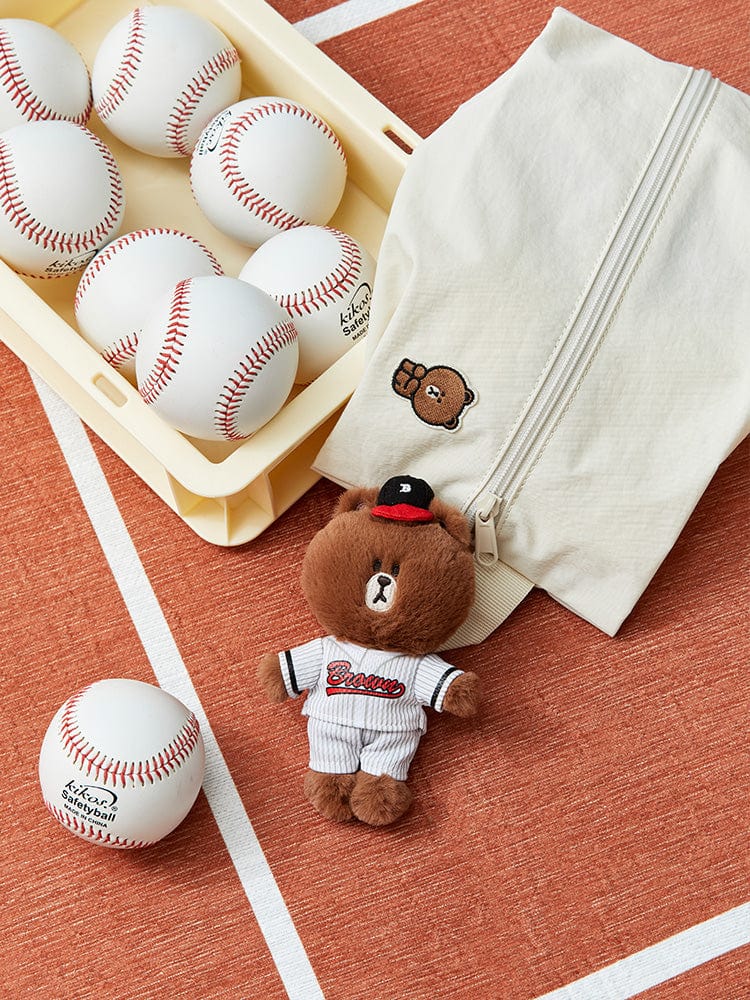 Close-up studio shot of a baseball-themed still life with baseballs in a beige tray, a beige zippered bag with a bear patch, and a stuffed brown bear in a baseball uniform on an orange and white striped surface.