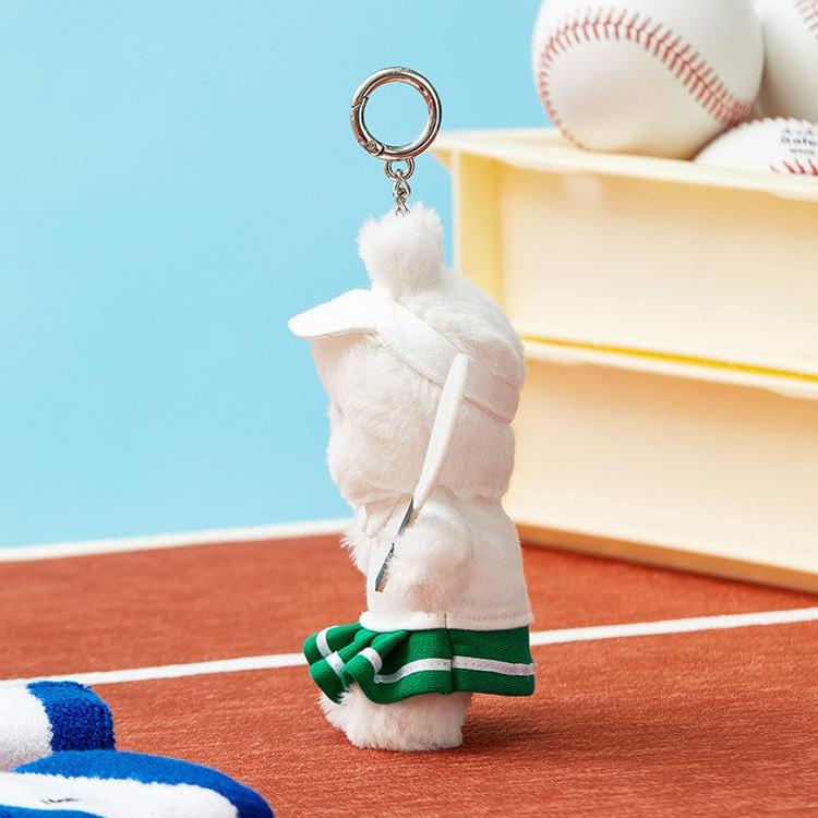 A plush, white rabbit keychain dressed in a tennis outfit with a green and white skirt, white visor, and a tennis racket, on a red surface with a blue backdrop.
