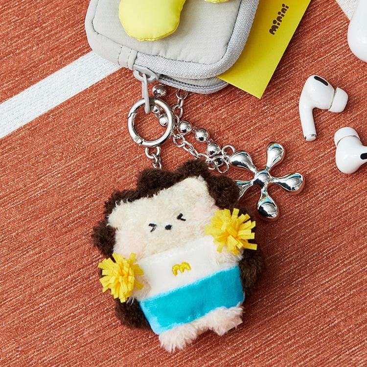 Plush keychain with brown fur, white face, blue and white outfit, and yellow pom-poms, attached to a silver chain, and white earphones on a textured orange surface.