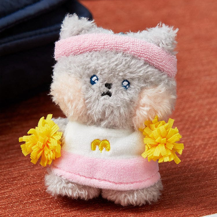 Gray plush toy cat with a pink headband, cheerleader outfit with a yellow "M" on the front, and yellow pom-poms.