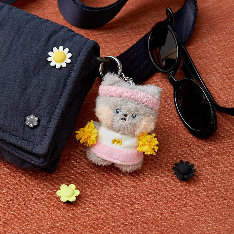 Product image of a dark blue bag with a white daisy accessory and a key chain of a furry gray cat with a pink sweatband and pink vest holding yellow pom poms, next to black sunglasses, and light green and black flower accessories on a textured orange surface.