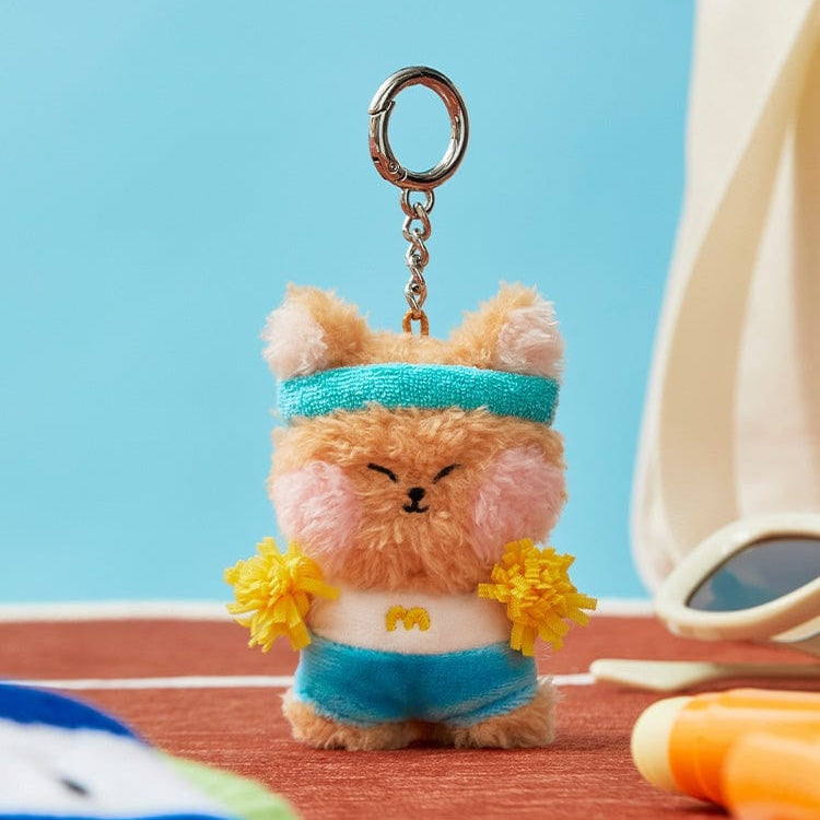 A fluffy, brown bear keychain with a blue headband, blue shorts, a white top with a yellow 'M', and yellow pom-poms.