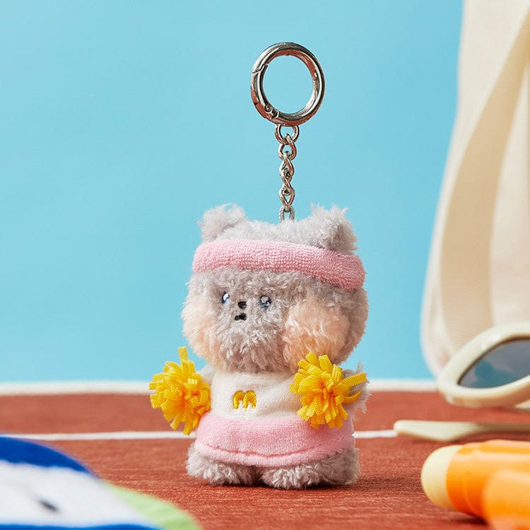 A plush grey hamster keychain wearing a pink headband, white shirt with a yellow 'M', pink skirt, and yellow pom-poms, sitting on a red track.