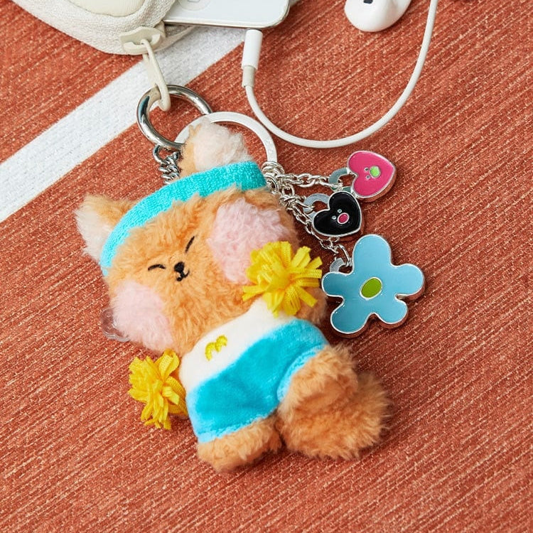 Close-up of a plush keychain with a tan fox character wearing a blue headband and shorts, attached to a silver ring with charms shaped like a heart, a black heart, and a blue flower.