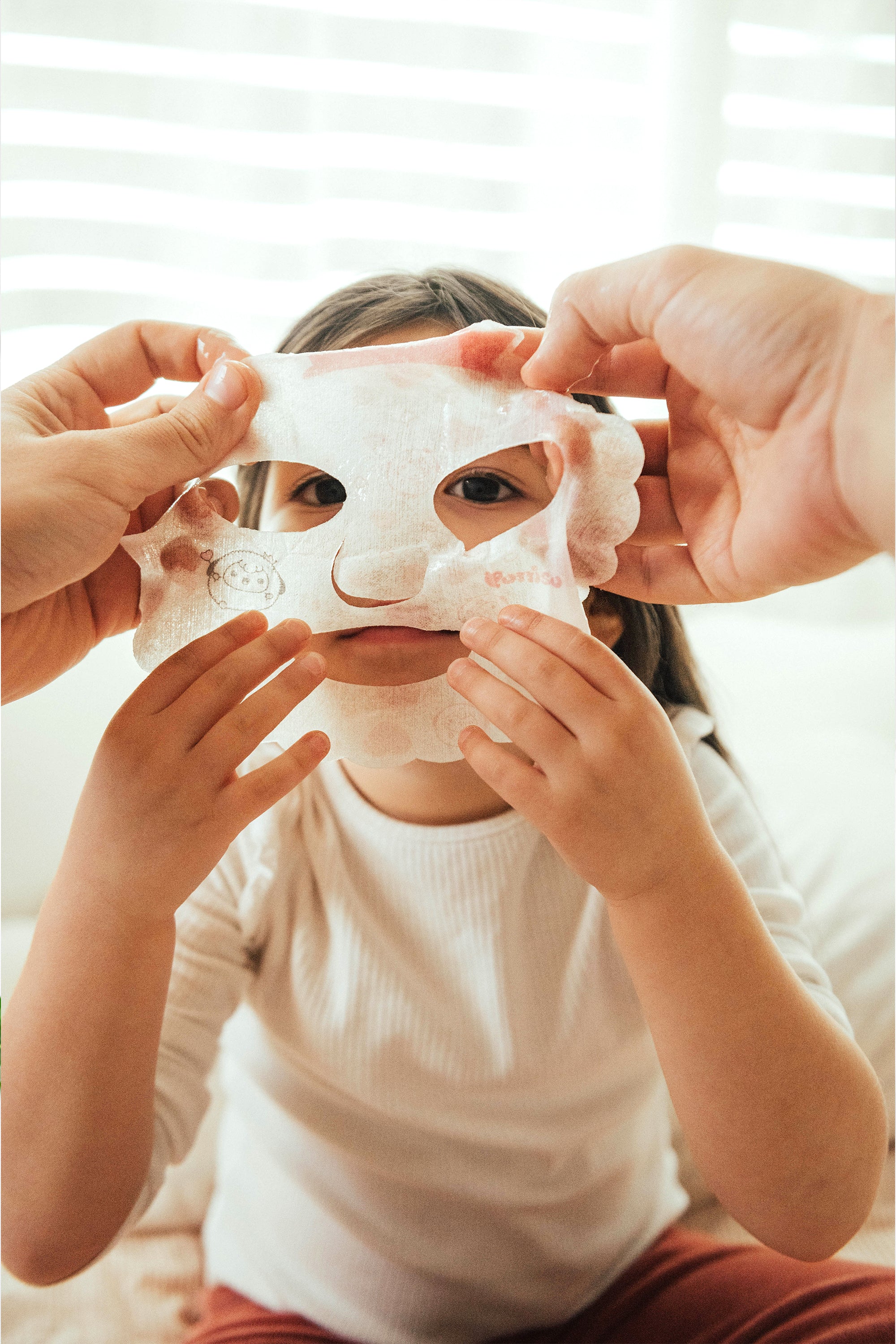 A child applying a face mask with assistance from adult hands.