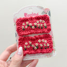 Two red, crocheted hair clips with embroidered white and yellow flowers on a floral patterned product card held in a hand.