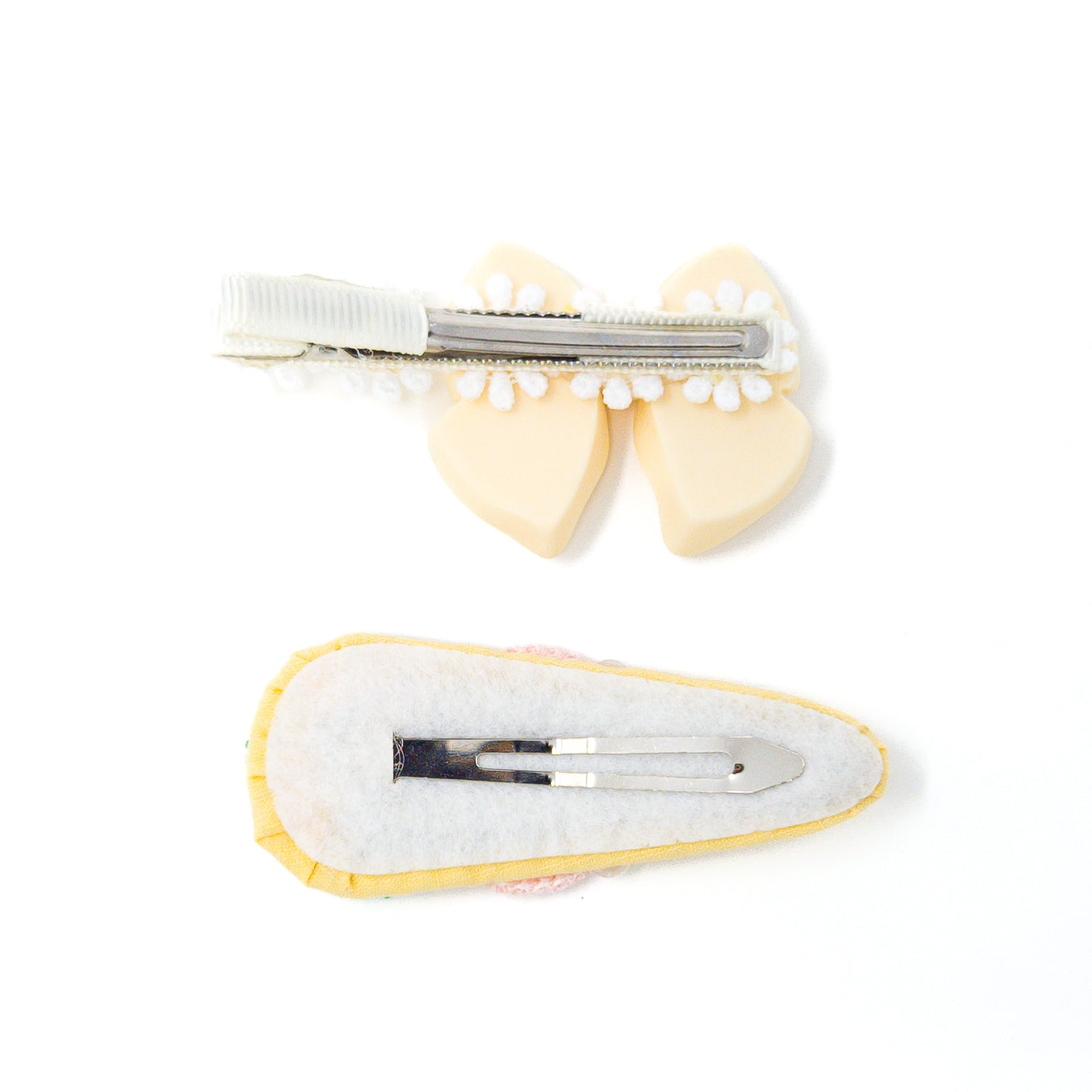 Two hair clips, one light yellow bow with a white and silver clip, and one pastel fabric clip with a silver clip. Both clips are against a white background.