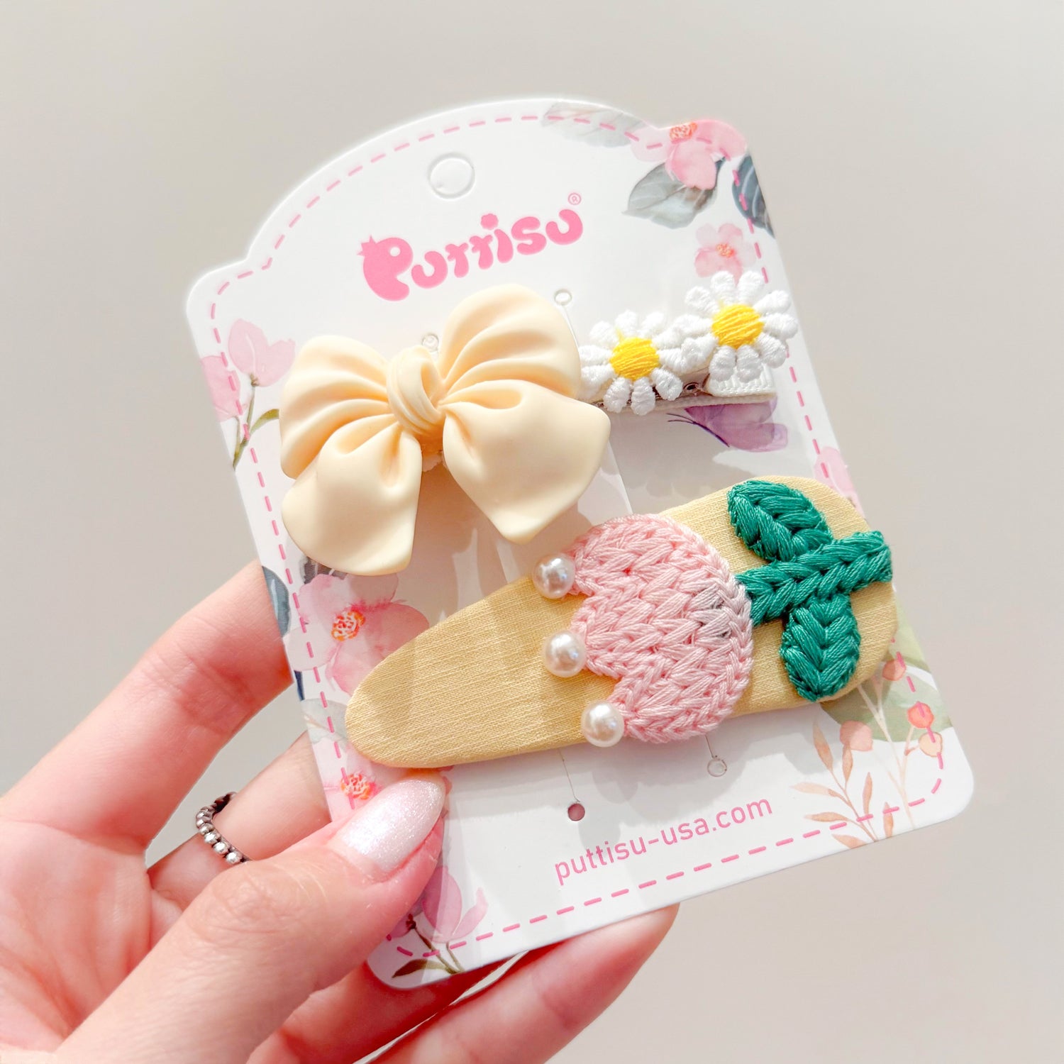 A set of three hair clips: a yellow bow, two daisy flowers, and a pink and green embroidered leaf shape with pearls, all displayed on a floral patterned card.