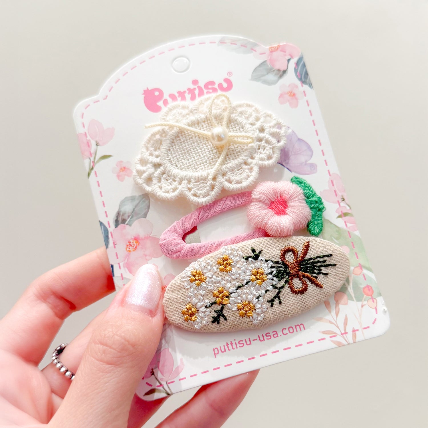 A hand holds a card of three hair clips. One clip is a white, lacy flower with a pearl center. The next is pink, with a pink flower and green leaf. The last is an oval of tan fabric embroidered with white and gold flowers, and a brown bow. The card is white with a floral pattern.