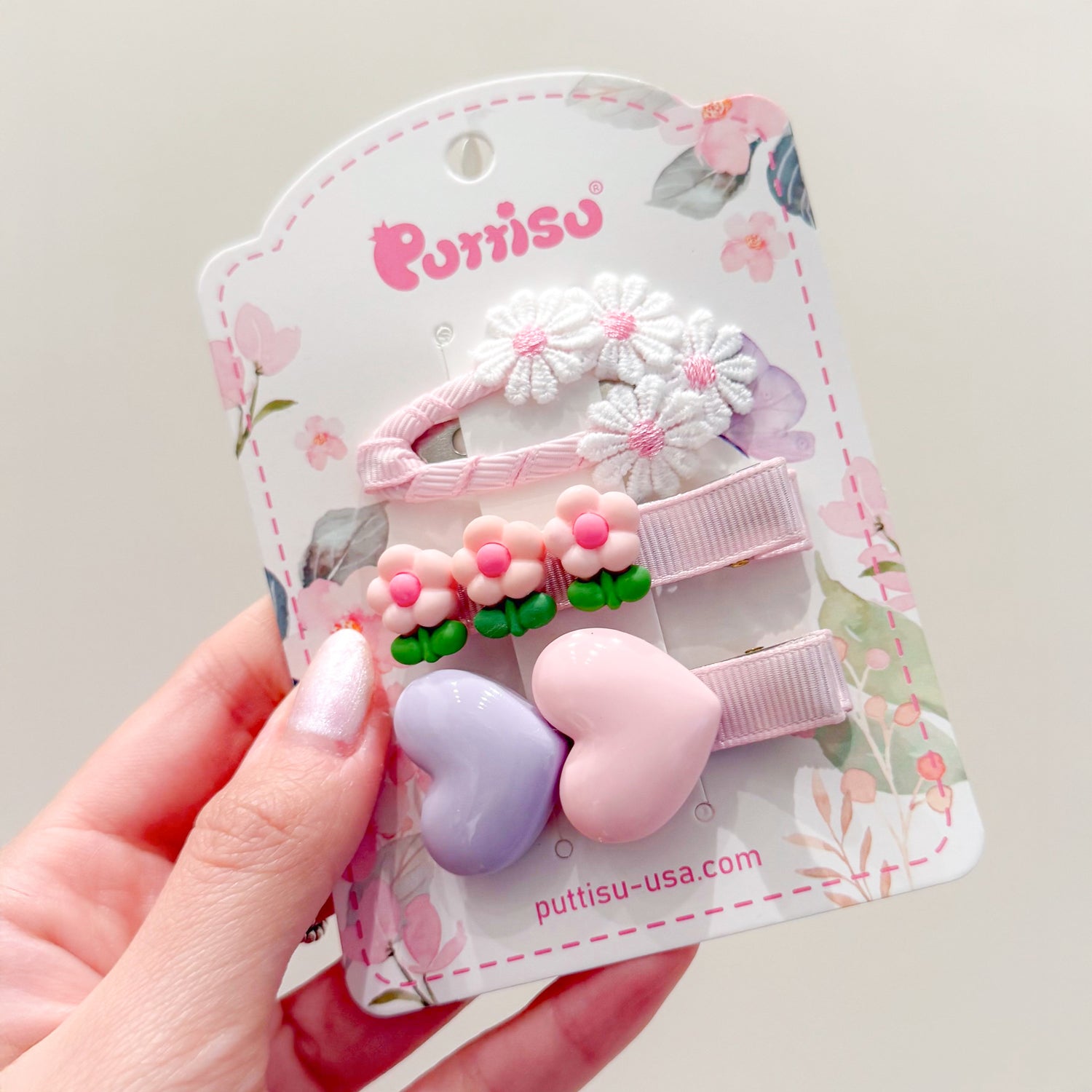 A hand holds a retail display card with three hair clips. The clips feature white daisies, pink flowers, and purple and pink hearts.