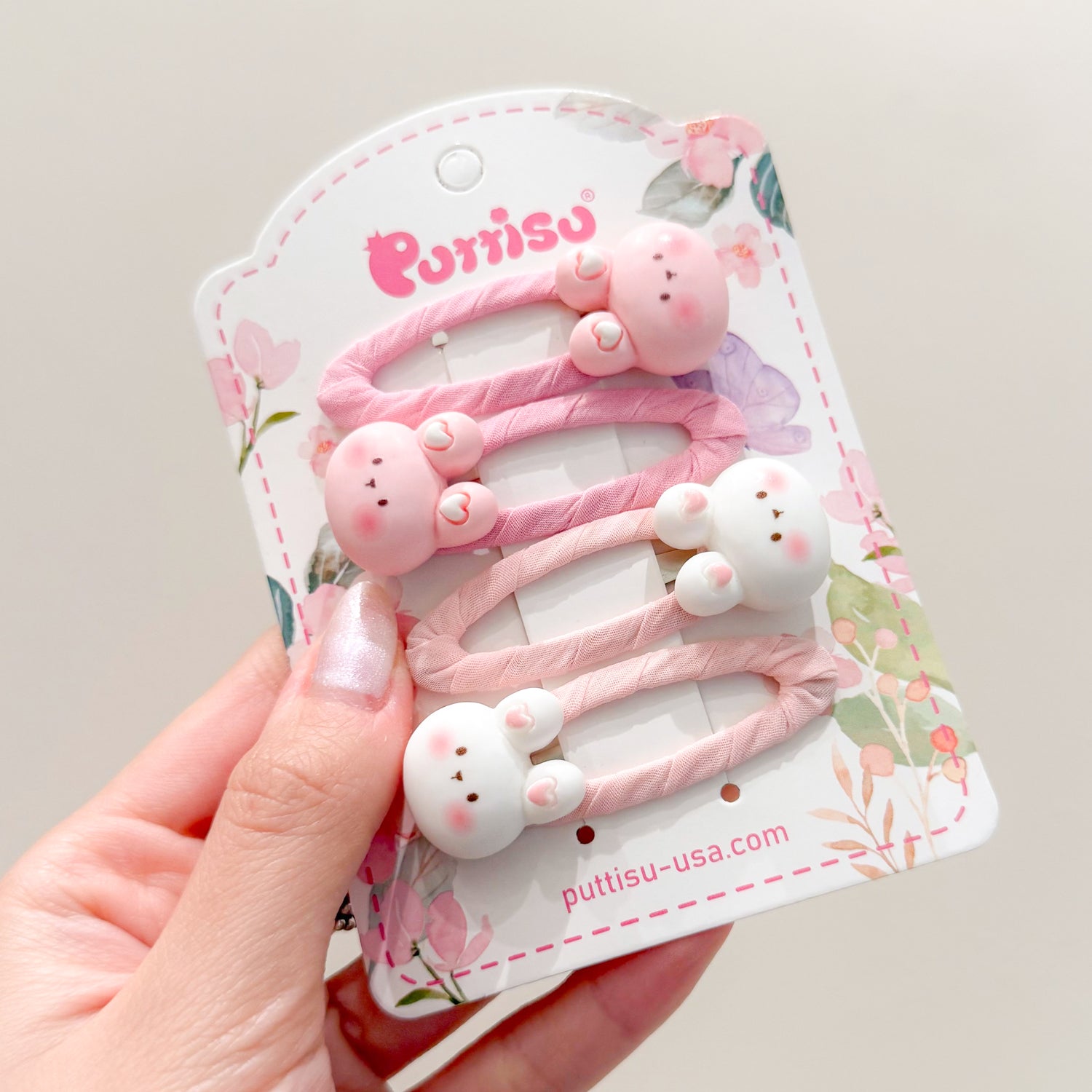 Three pink and white bunny hair clips on a floral-themed product card held by a hand.