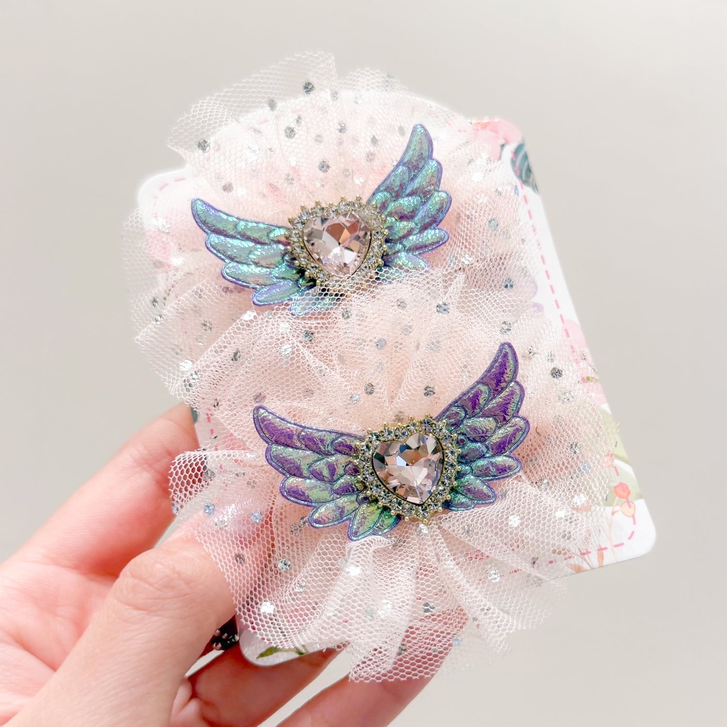 Close-up of two hair clips with heart-shaped jewels with wings on a pink frilly, sparkly netting. One is blue, one is purple. The hairclips are being held up by someone's hand.