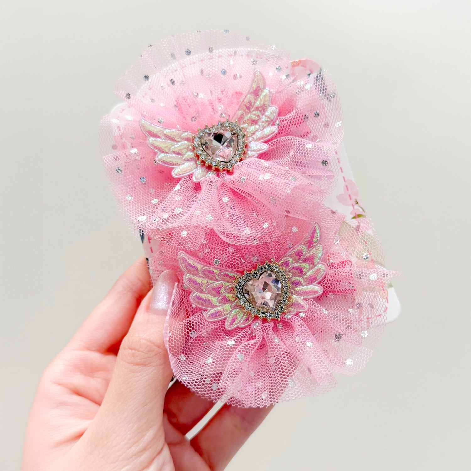 Close-up of a hand holding two pink, sparkly hair clips with heart-shaped rhinestones and wings.