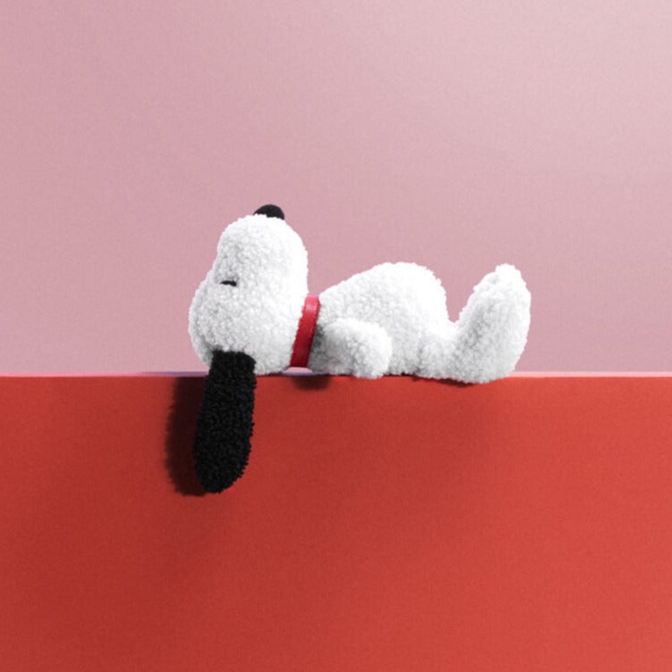 Stuffed Snoopy dog with a red collar, lying on a red surface, against a pink background.