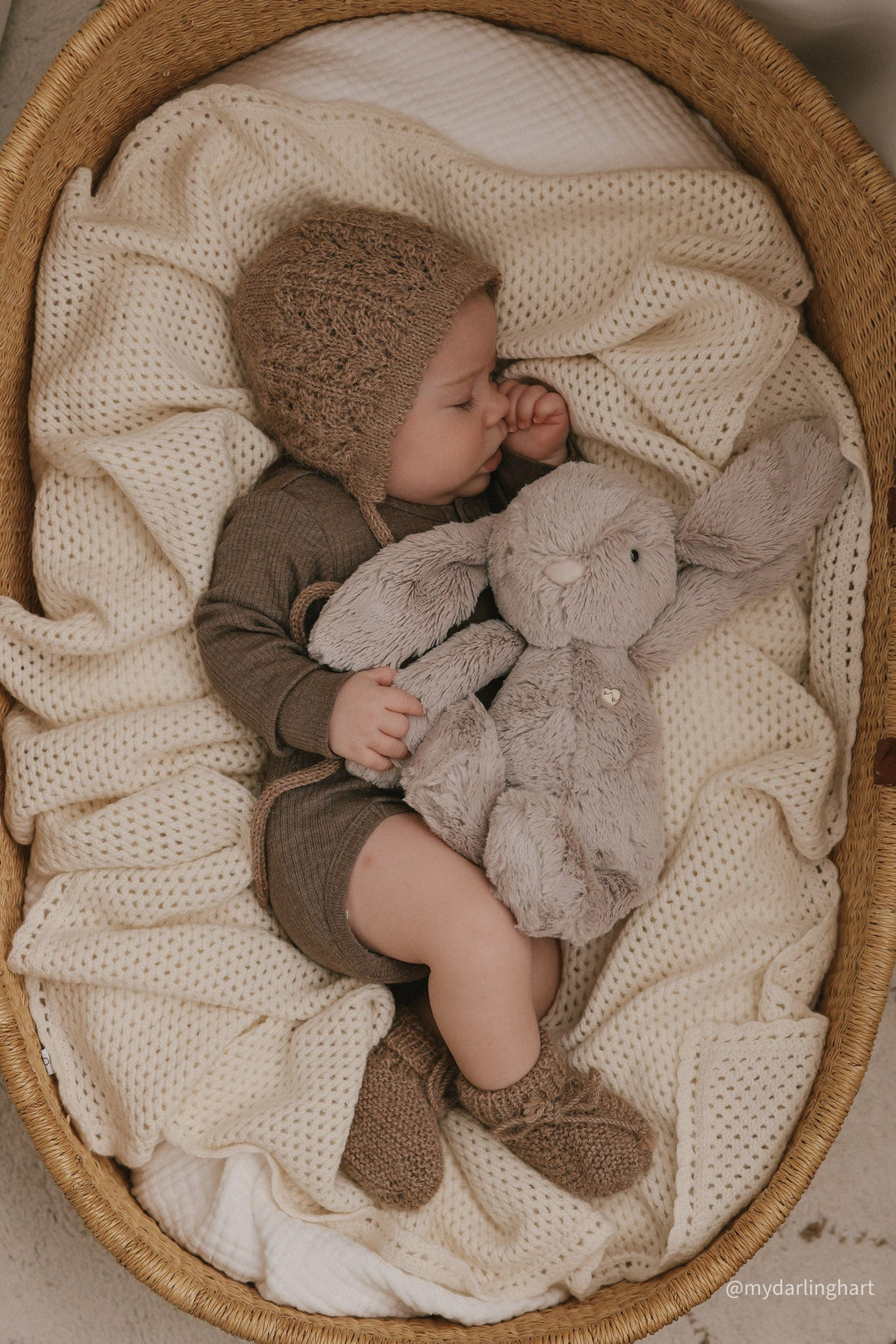 Baby sleeping in a bassinet with a stuffed bunny, wearing a knitted bonnet and socks.