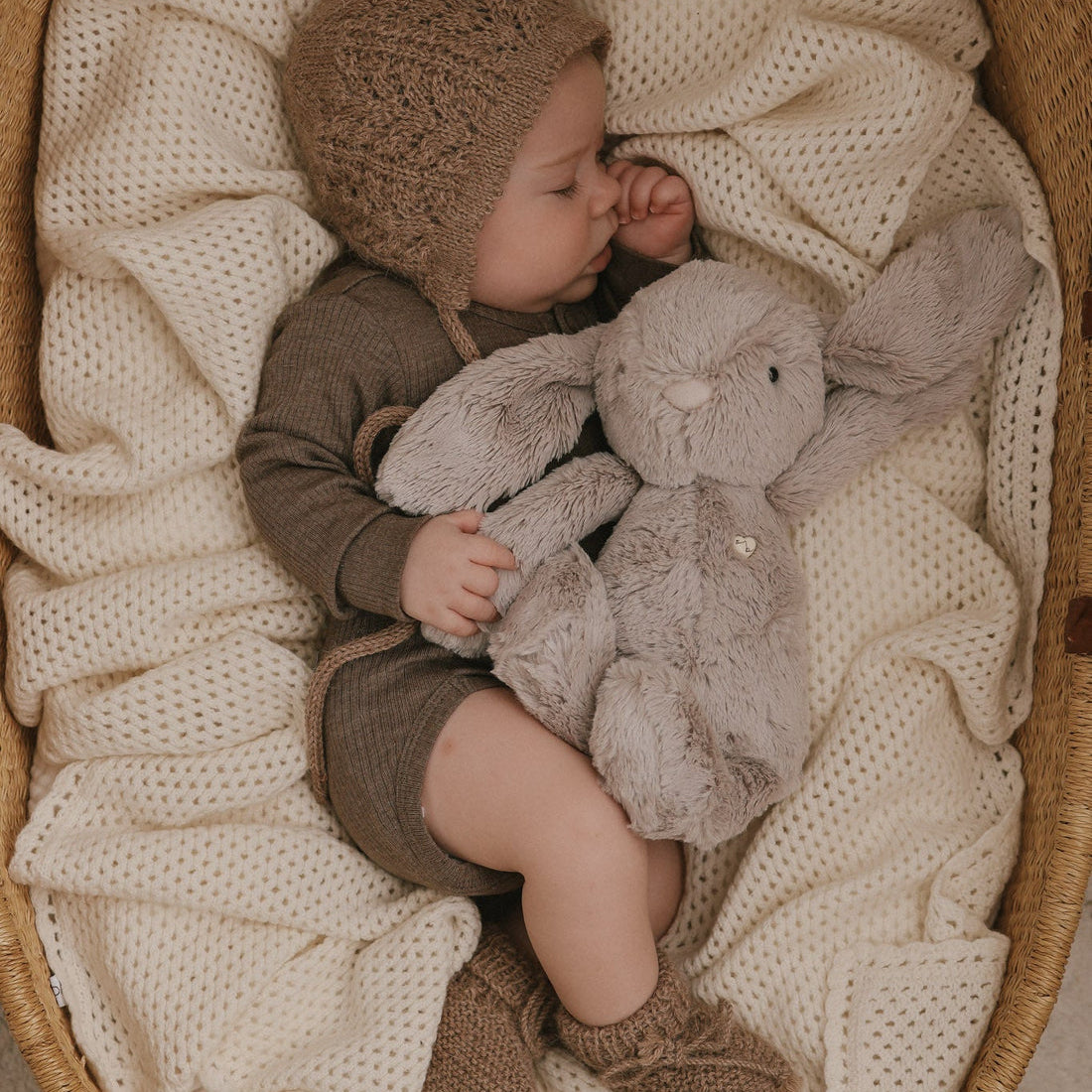 Baby sleeping in a bassinet with a stuffed bunny, wearing a knitted bonnet and socks.