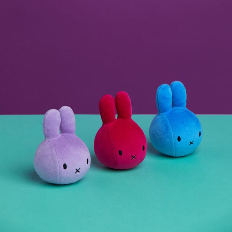 Three plush bunny toys in purple, red, and blue on a teal surface against a purple background.