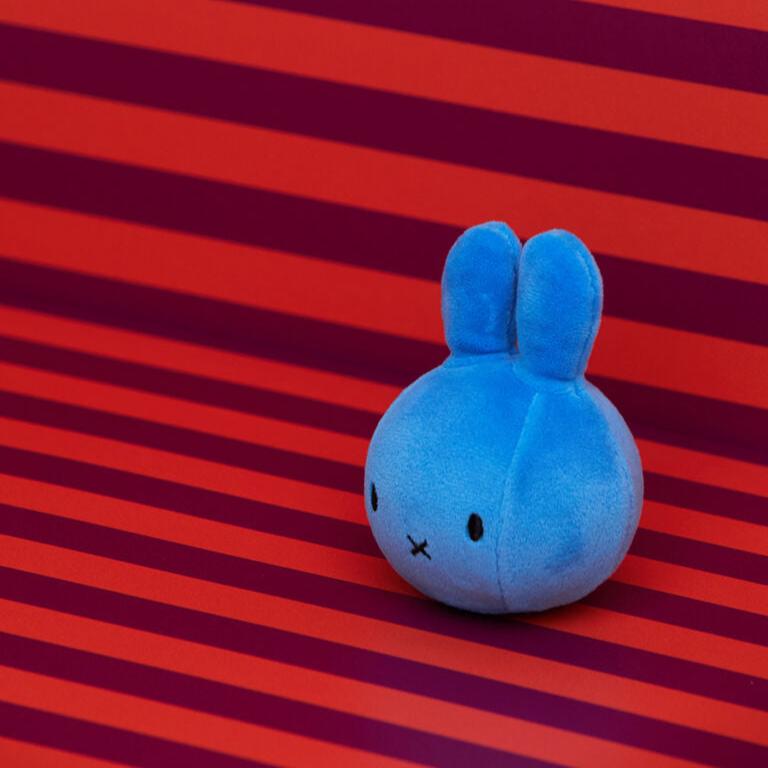 A blue Miffy plush toy against a striped red background.