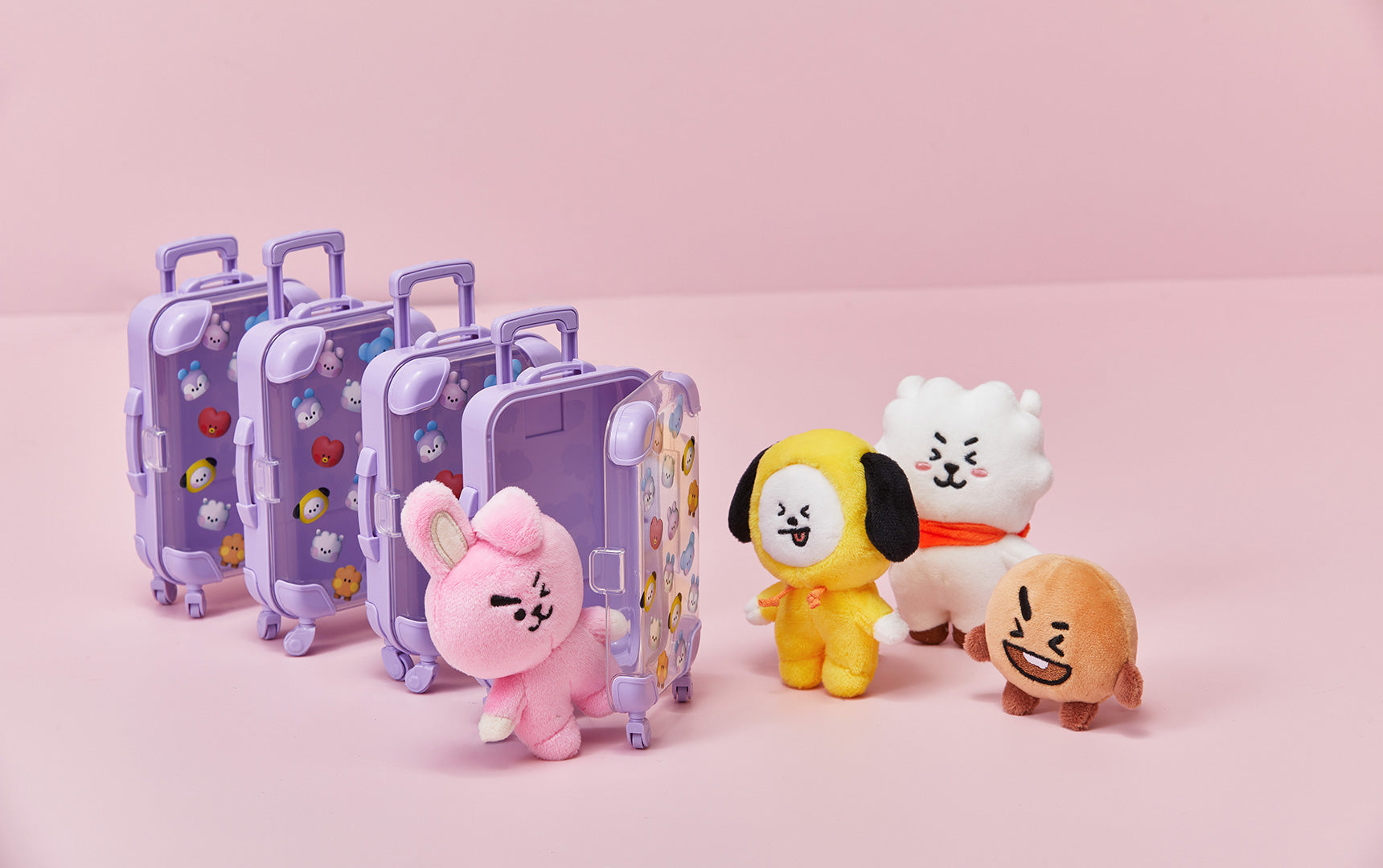 Miniature purple toy suitcases with character stickers and small stuffed BT21 plush dolls on a pink surface.