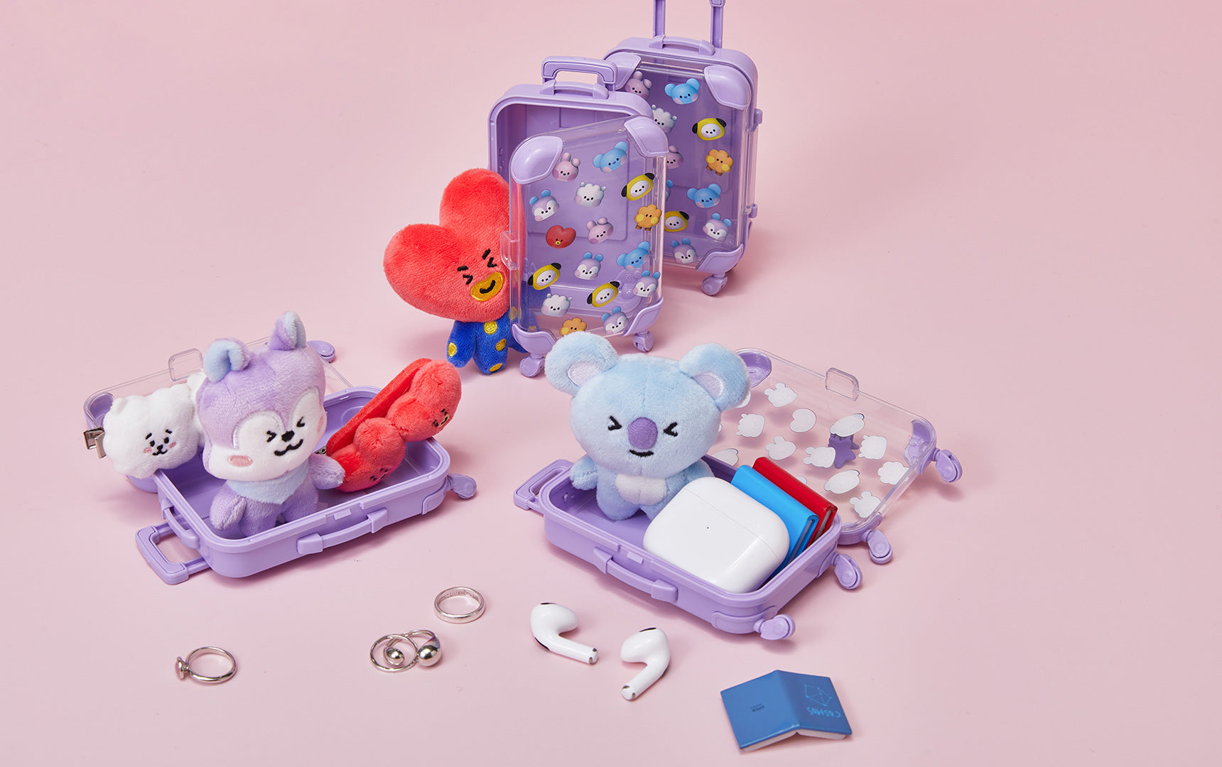 Still life featuring purple toy suitcases, BT21 plush toys, AirPods, and various rings on a light pink background.