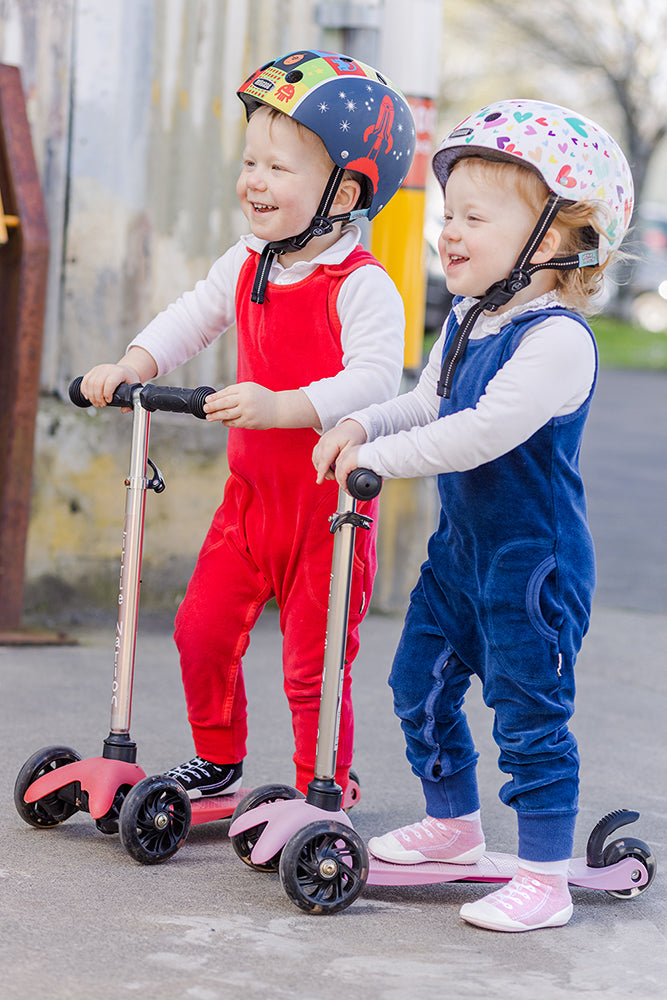 Two young children in helmets and brightly colored jumpsuits ride scooters on a paved surface.