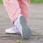 Close-up of child's feet wearing pink corduroy pants, white socks, and lavender Komuello shoes with grey soles on a brick walkway.