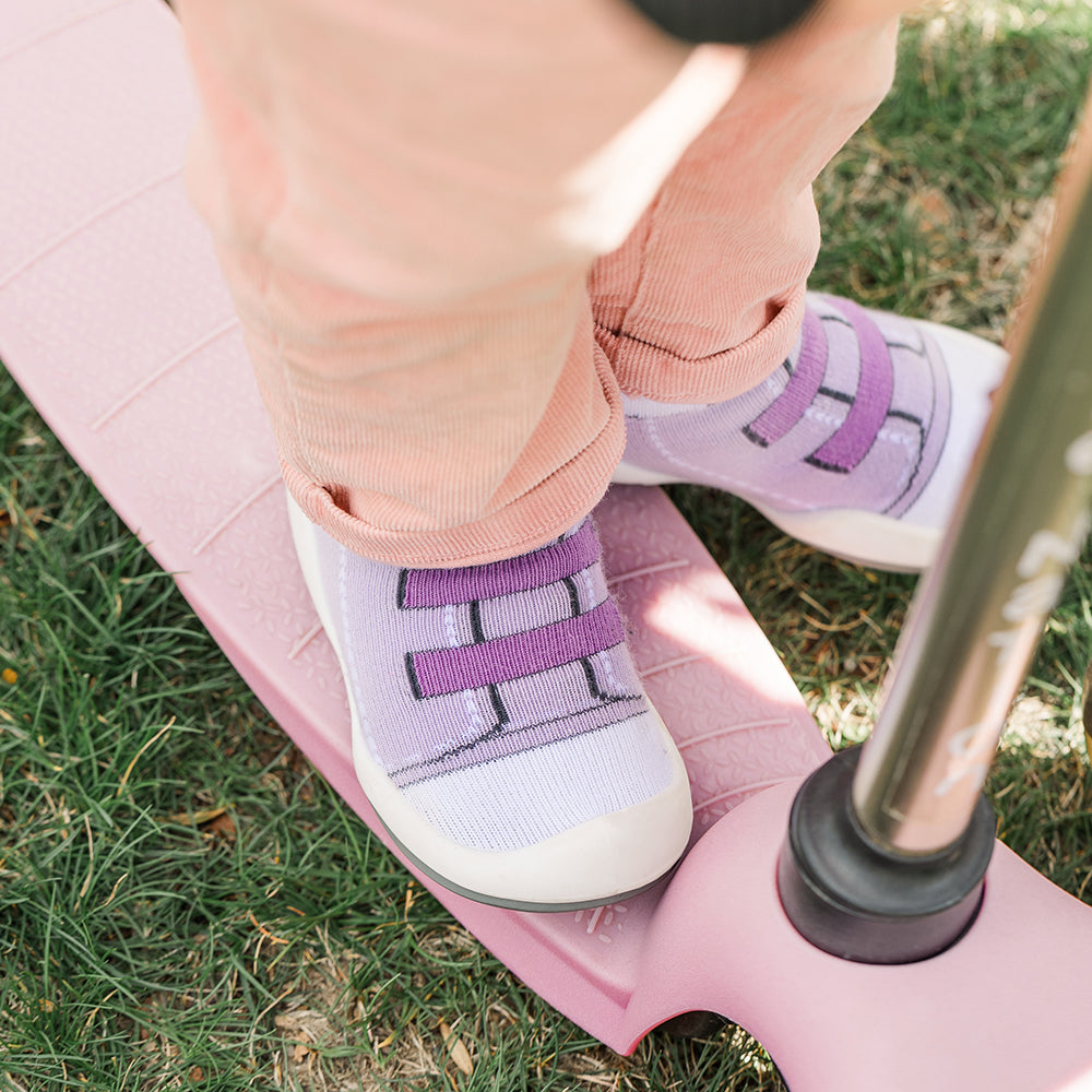 Child in pink pants and lavender sneakers stands on a pink scooter on grass.