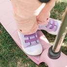 Child in pink pants and lavender sneakers stands on a pink scooter on grass.