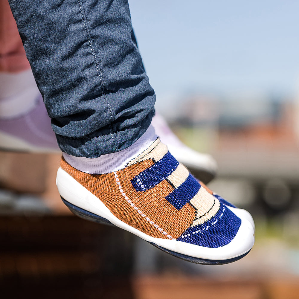 A child's foot wearing a brown and blue sock shoe with a white sole.