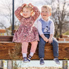 Two young children sitting on a wooden beam, girl in a floral dress and pink leggings, boy in a blue sweater vest and jeans.