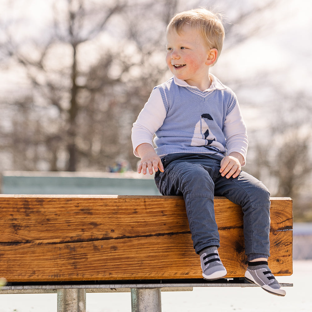 Young boy in blue sweater vest and pants sitting on wooden beam outdoors.