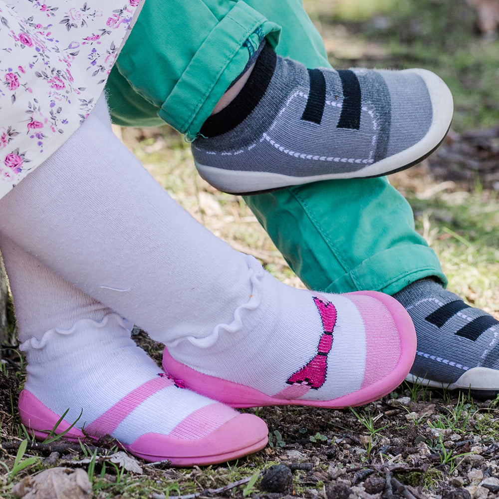 Two children's legs, one wearing pink and white striped sock shoes with a bow design, the other wearing gray and black sock shoes with green pants.