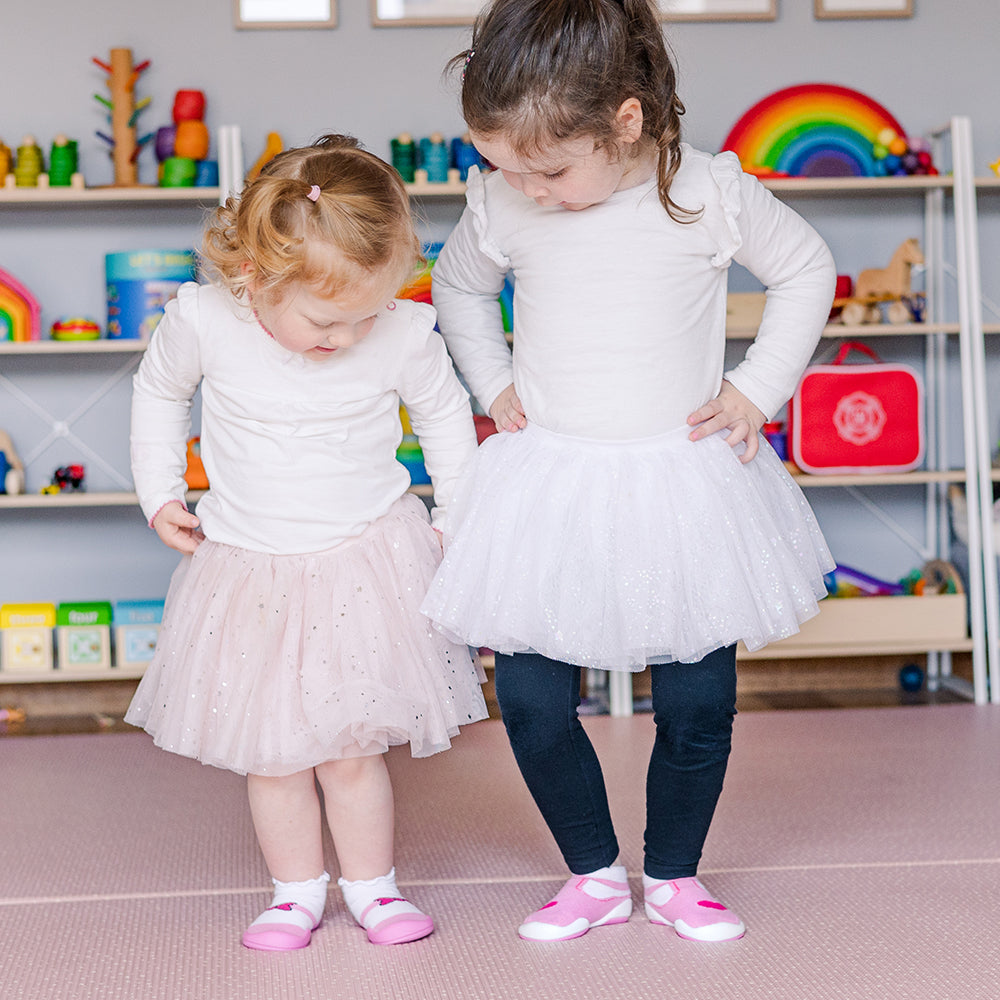 Two young girls in white long-sleeved shirts and tutus, one pink and one white, wearing pink and white shoes in a playroom.