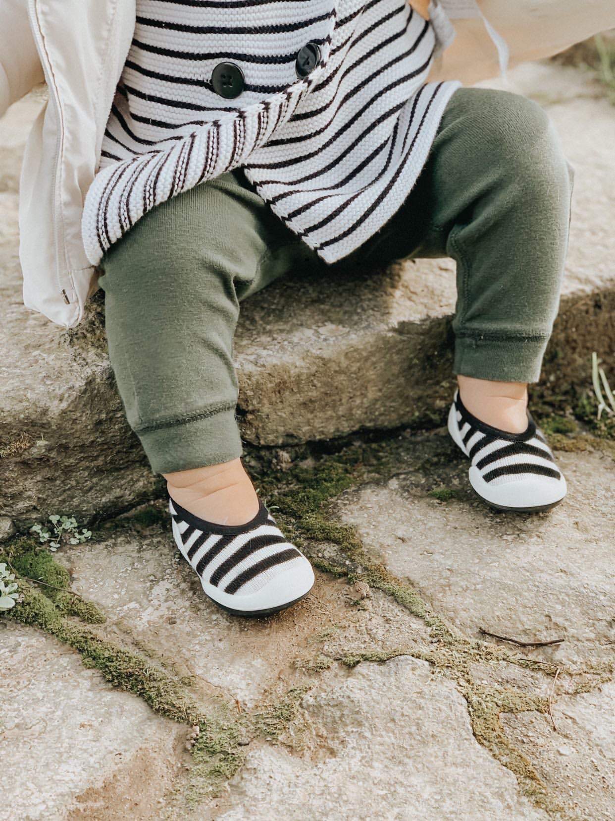 Baby wearing striped shirt, green pants, and striped shoes sitting on stone steps.