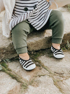 Baby wearing striped shirt, green pants, and striped shoes sitting on stone steps.