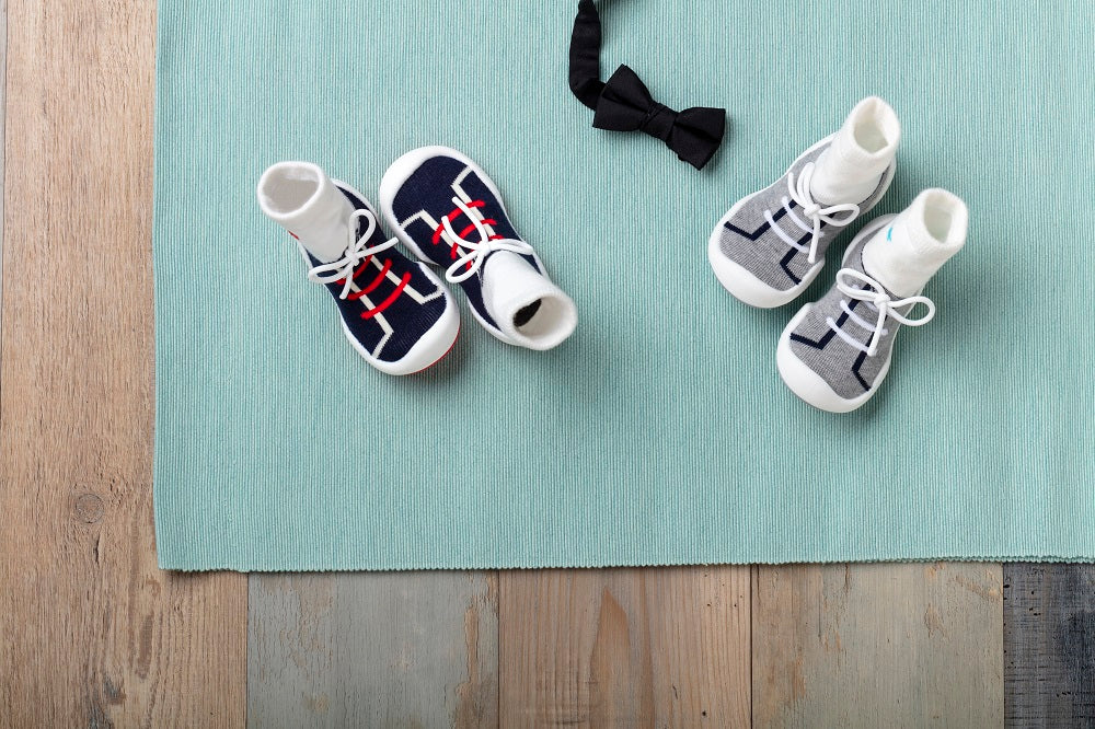 Two pairs of baby sneaker socks and a black bow tie on a turquoise mat, placed on a wooden surface.