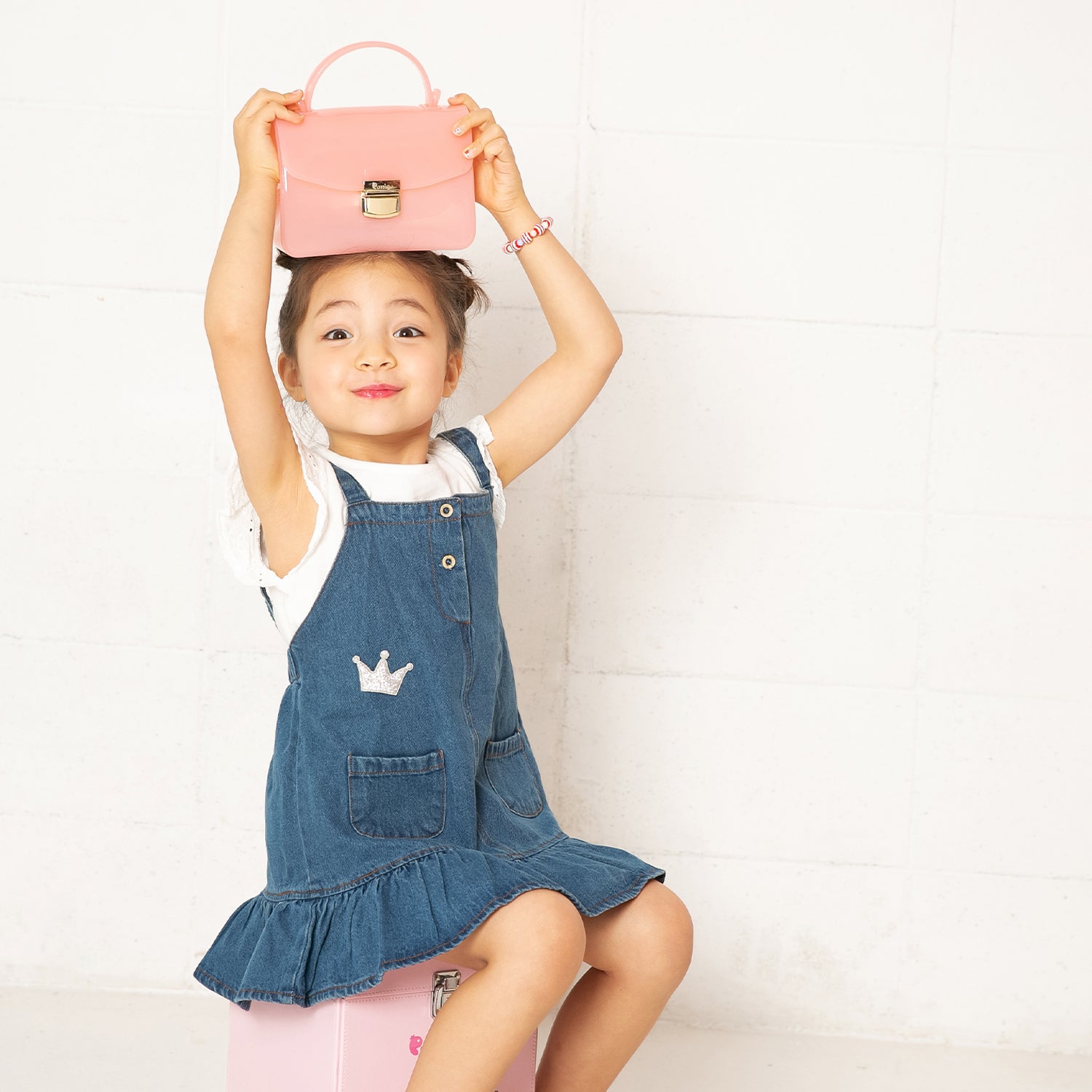 Girl in denim dress, seated with pink purse on head.