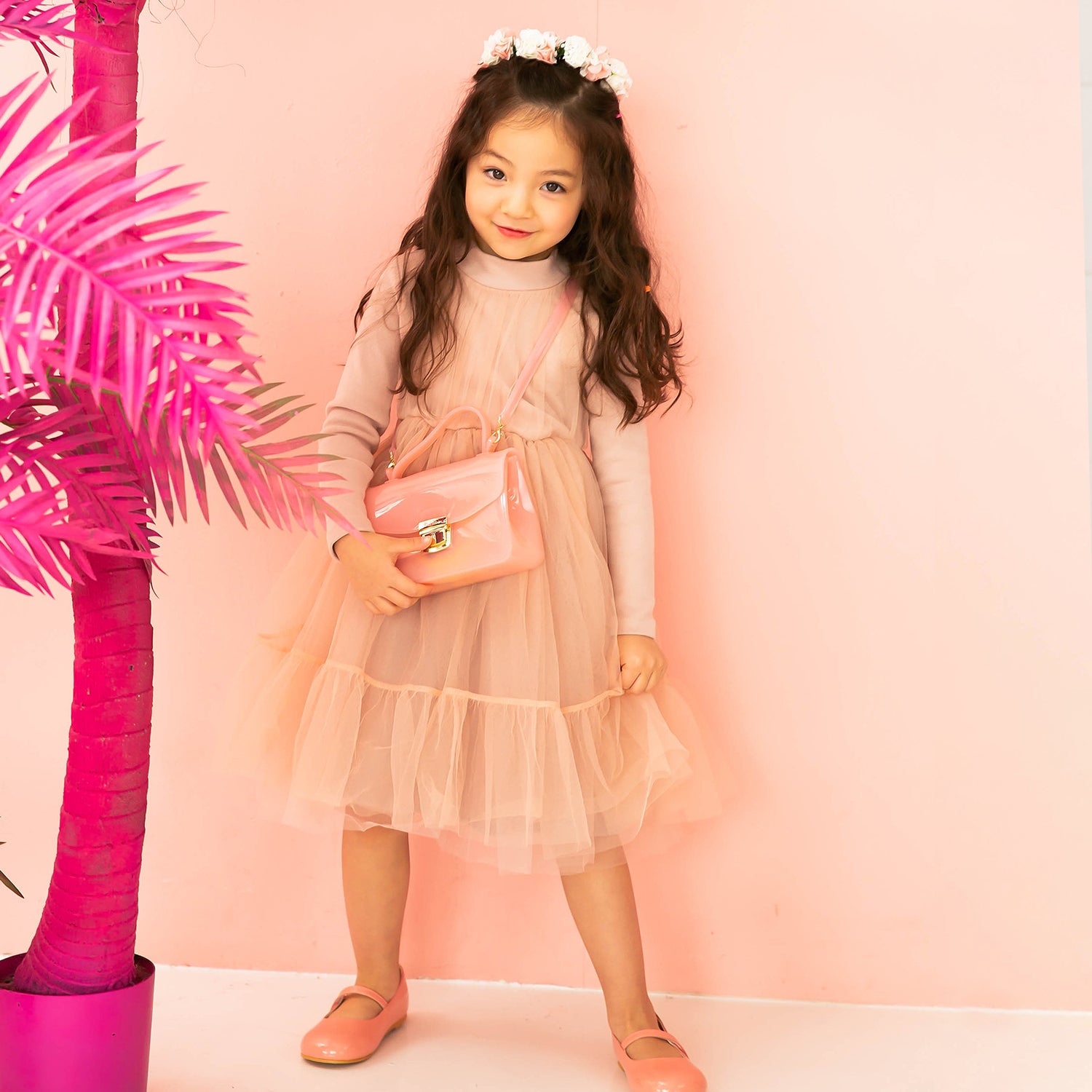 Young girl in a peach dress with a purse and flower crown posing in front of a pink palm tree.