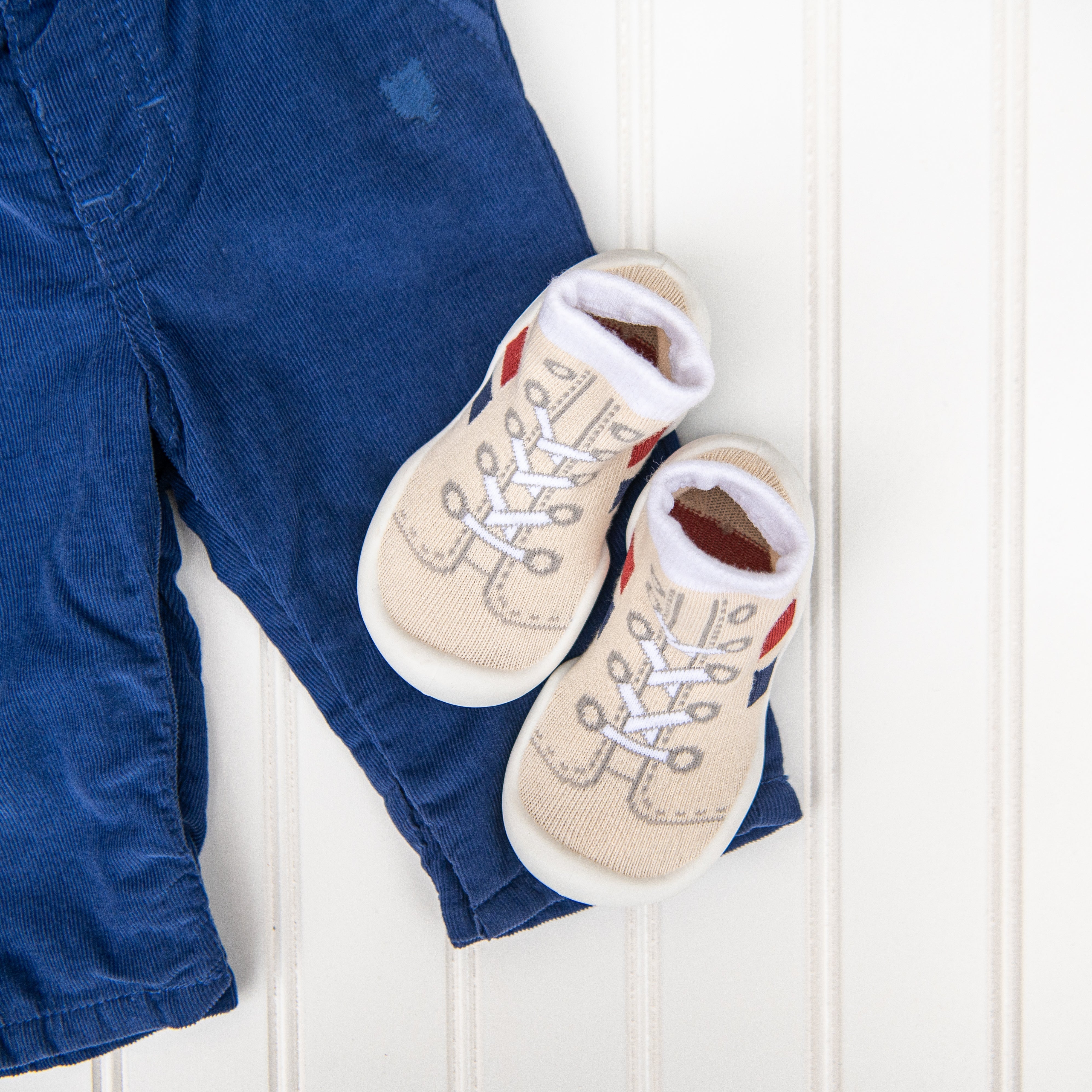 Top-down shot of blue corduroy pants with beige baby shoes that look like sneakers on top of the pants, placed on a white paneled surface.