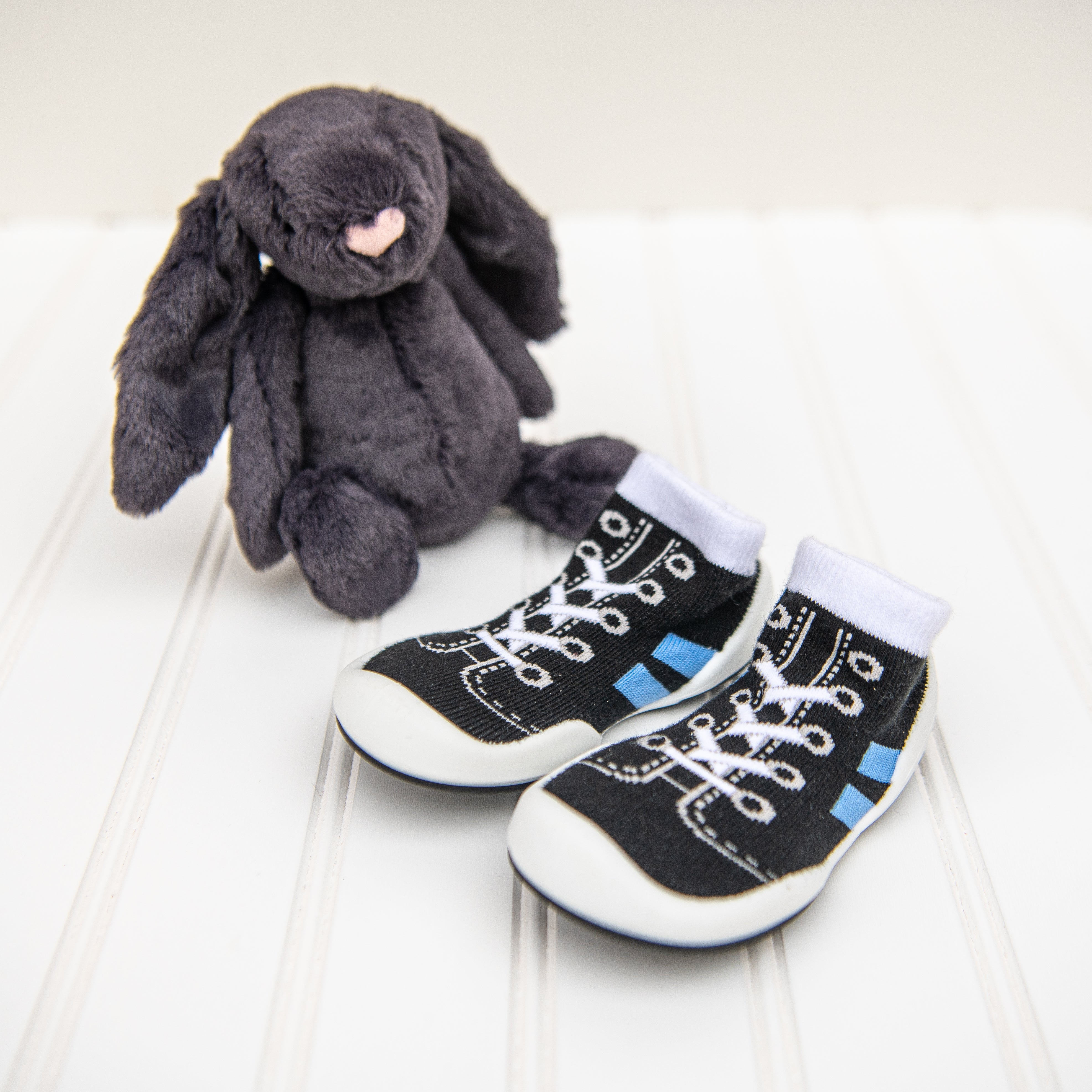 Pair of black baby shoe socks with white laces and blue stripes next to a gray plush bunny toy on a white wood surface.