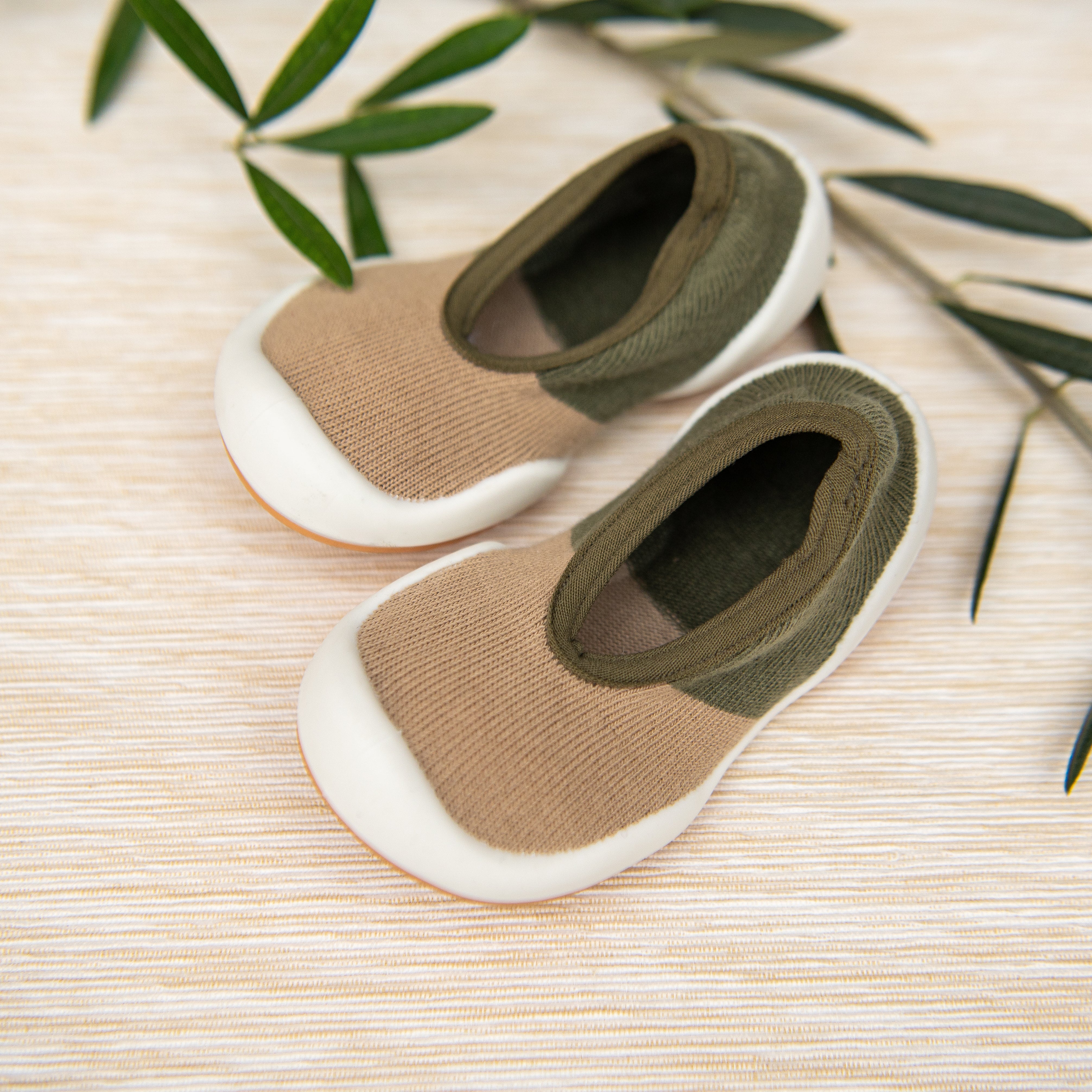 Pair of tan and green baby slip-on shoes on a light textured surface with green leaves.