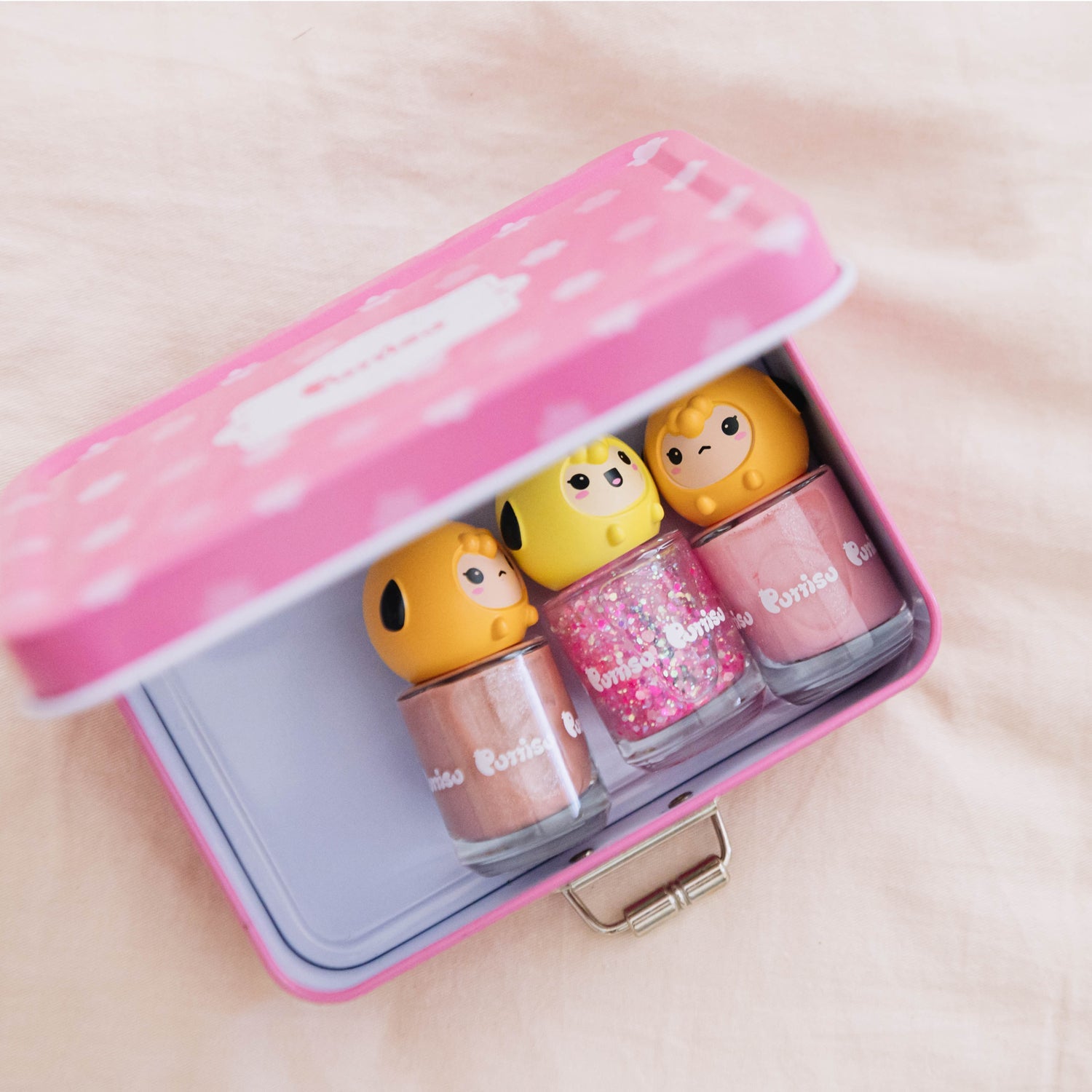 Close-up of a pink tin carrying three bottles of Miss Poniso nail polish. The bottles have yellow character faces on top, and contain light brown, pink glitter, and pink nail polish.