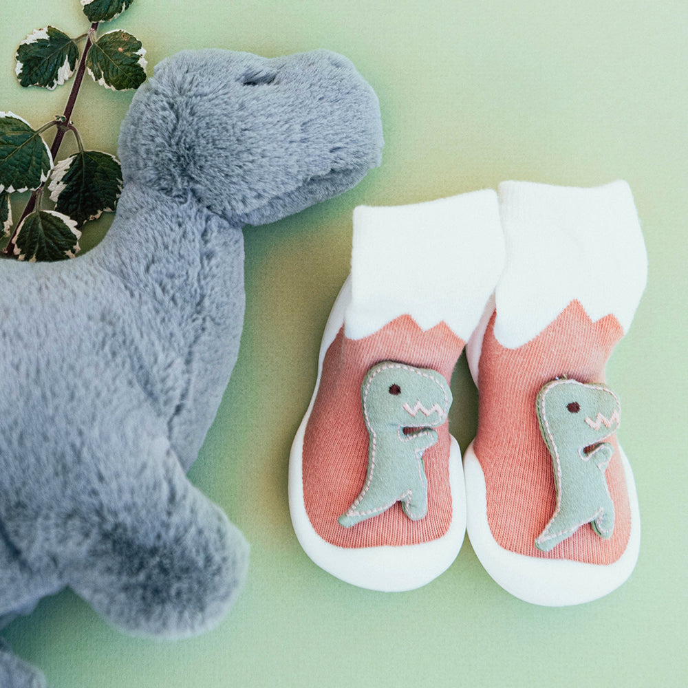 Pair of dinosaur-themed baby slipper socks with gray plush dinosaur toy.