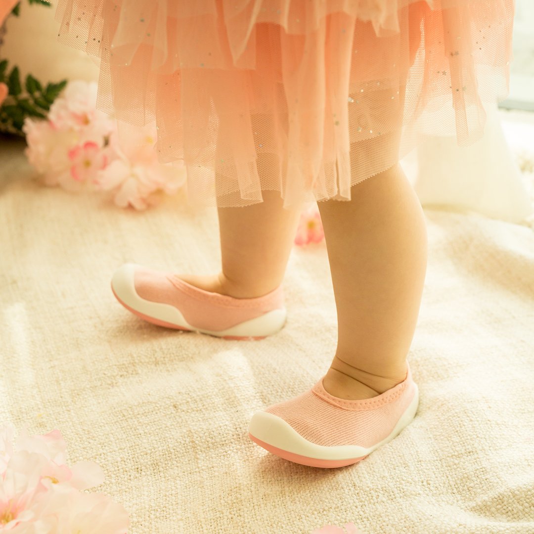 A baby or toddler wearing pink shoes and a pink dress stands on a cream-colored surface.