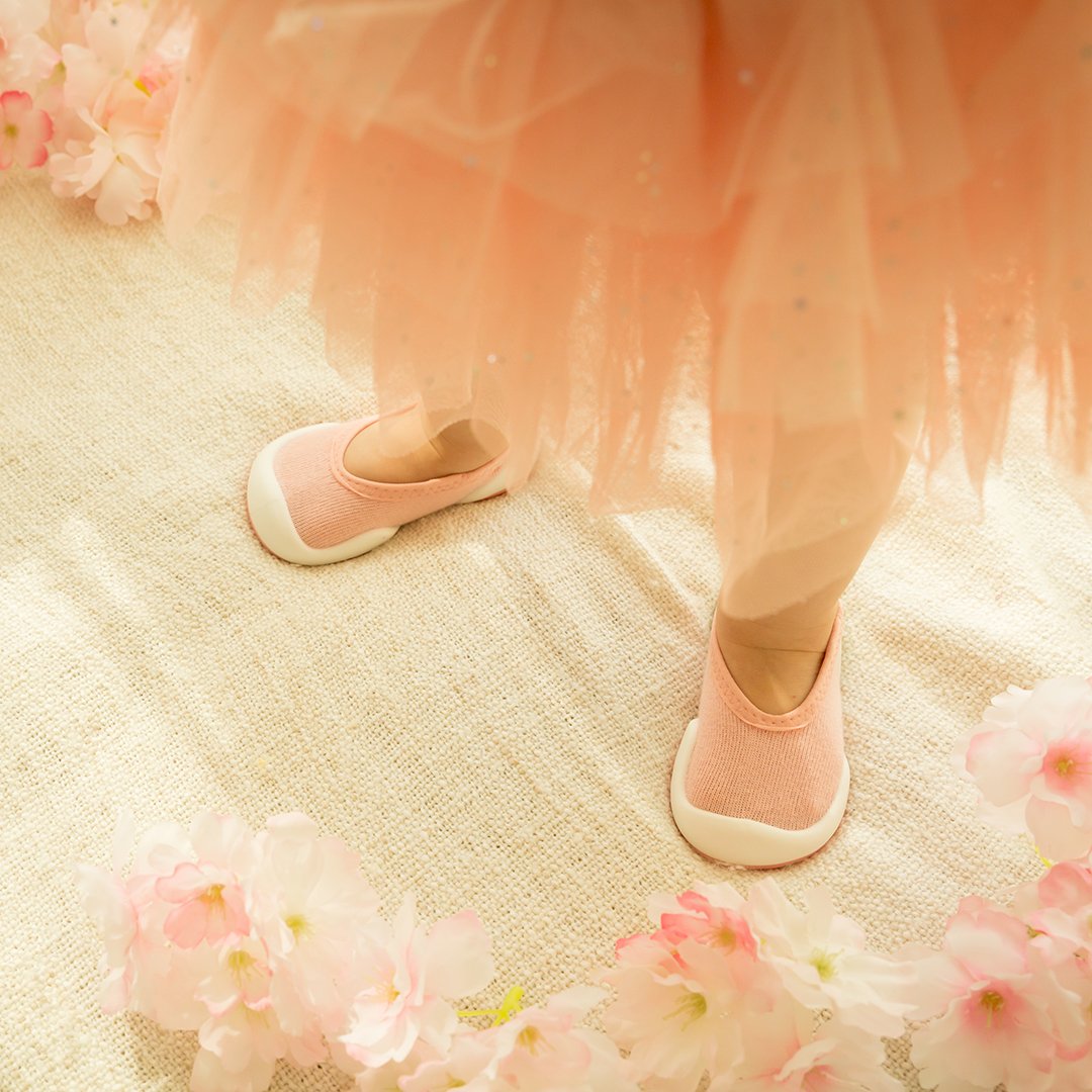 A child wearing pink shoes and a matching pink tutu standing on a textured surface surrounded by pink blossoms.