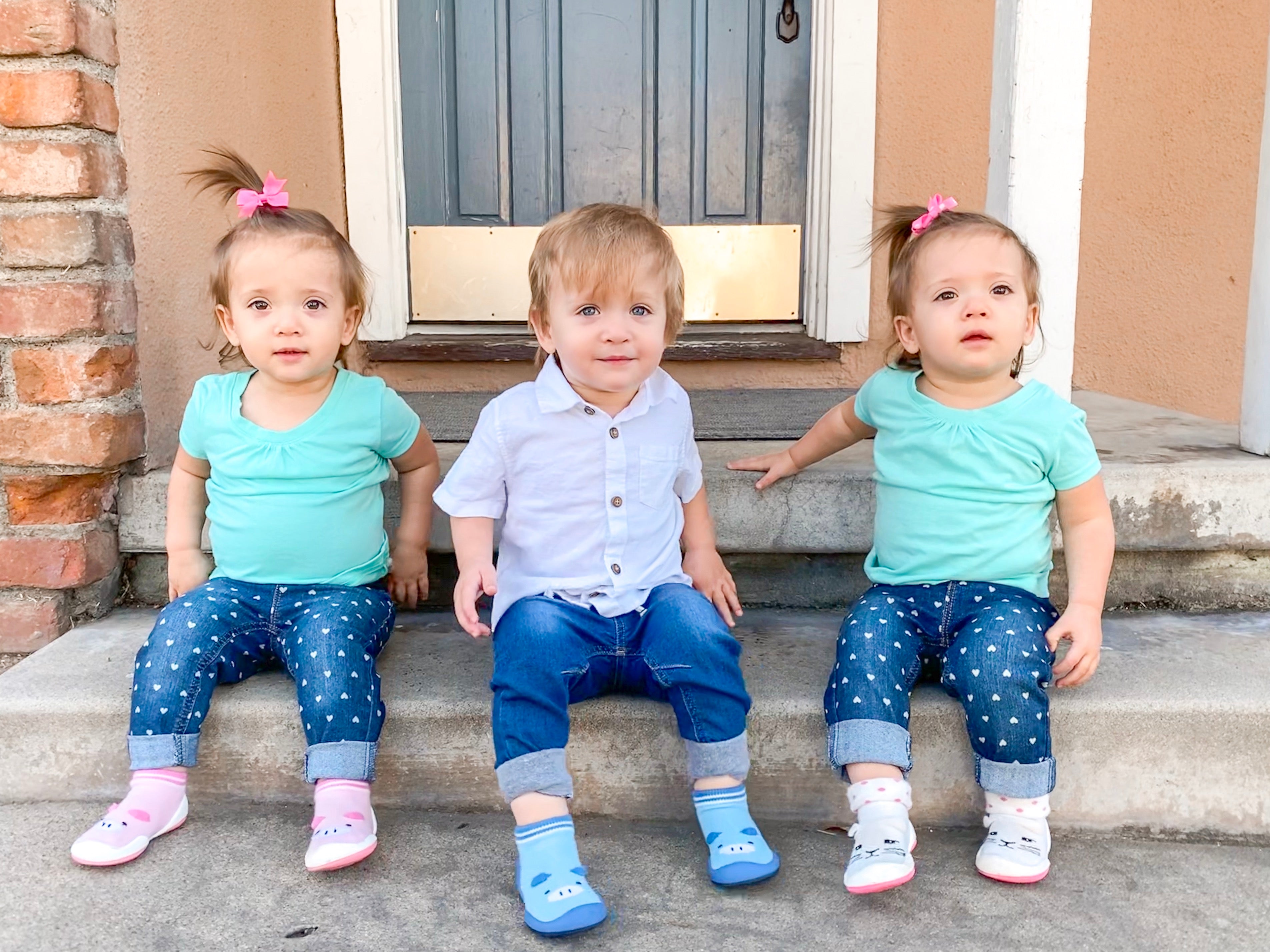 Three young children sit on stone steps. The two girls wear teal shirts, heart-patterned jeans, and animal-themed socks and shoes. The boy wears a white button-down shirt and blue jeans with matching socks and shoes.