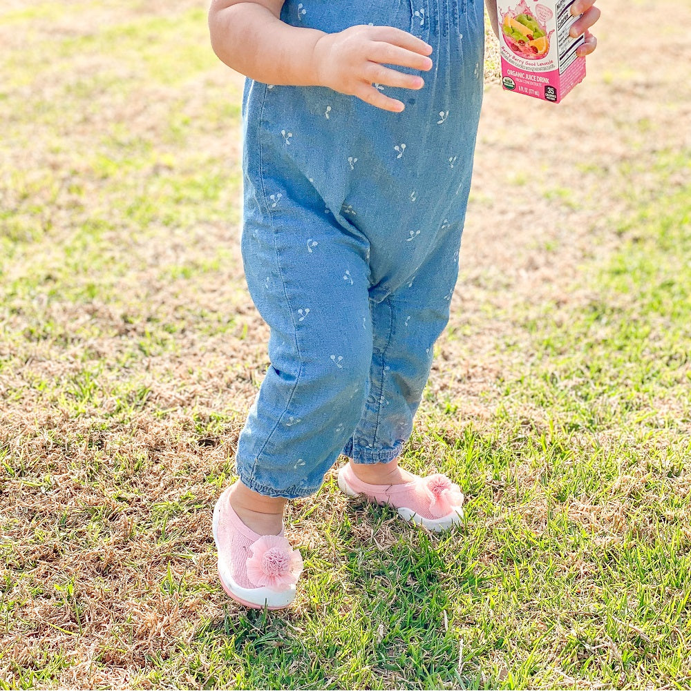 A baby in a denim jumpsuit and pink flower shoes stands on grass, holding a juice box.
